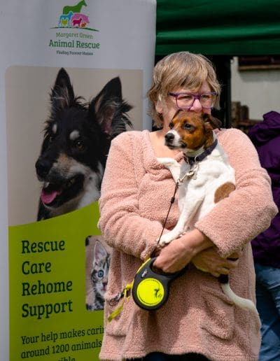 A woman is holding a brown and white Terrier in their arms, while stood in front of a promotional charity banner
