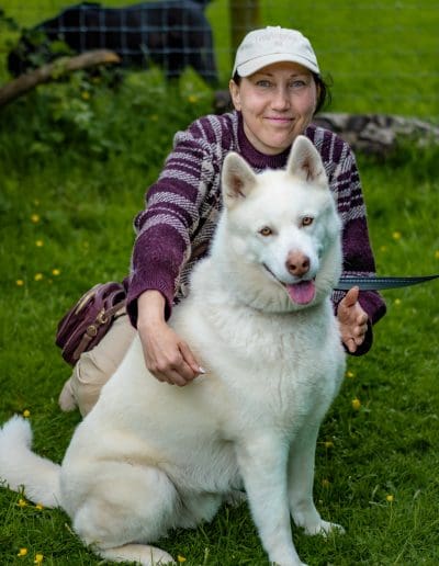 A woman is sat on the grass next to a white Husky and they are both looking at the camera